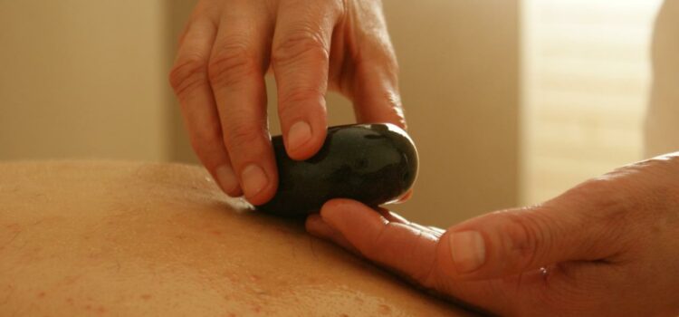 Le ou la massothérapeute dépose délicatement une pierre à massage sur le dos bien hydraté de la personne qui reçoit le soin